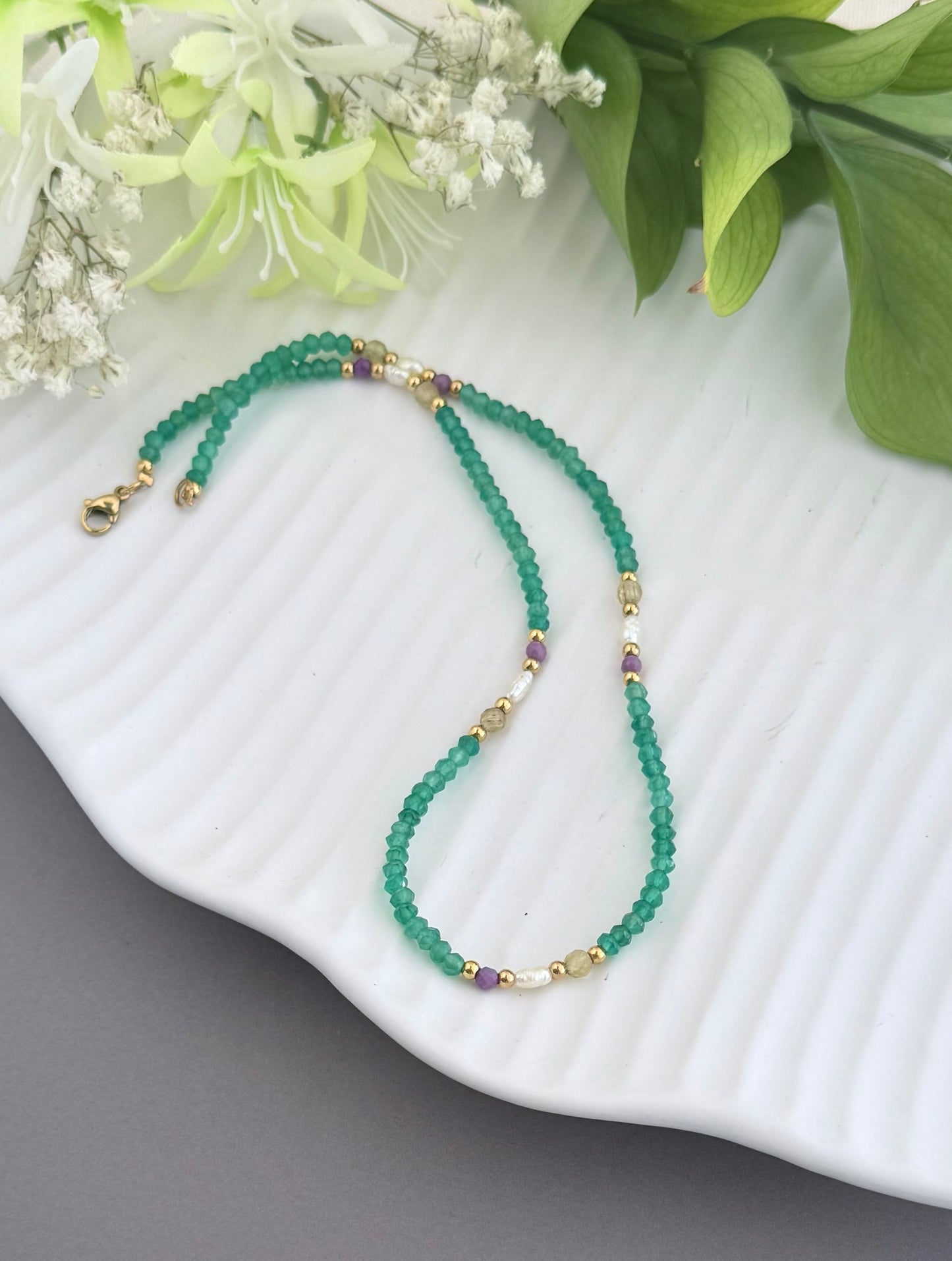 A beaded necklace made of green faceted beads with small gold spacer beads and a few purple accent beads, displayed on a white ribbed plate with green leaves and white flowers in the background.