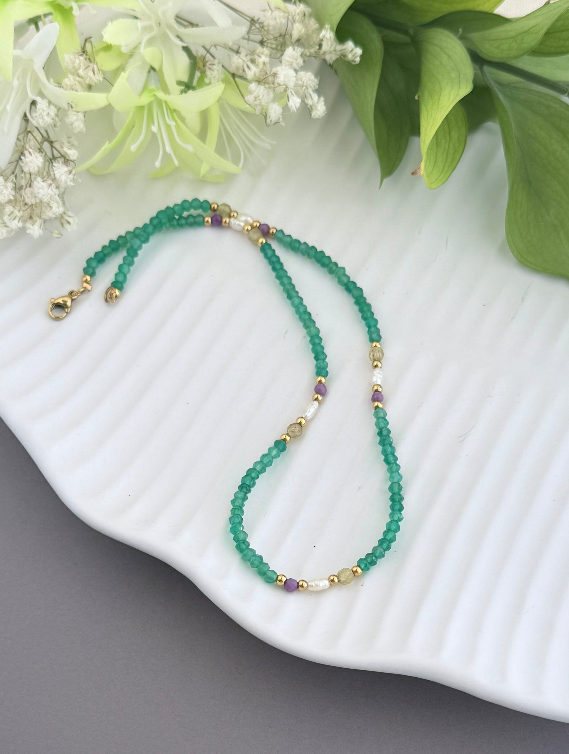 A beaded necklace made of green faceted beads with small gold spacer beads and a few purple accent beads, displayed on a white ribbed plate with green leaves and white flowers in the background.